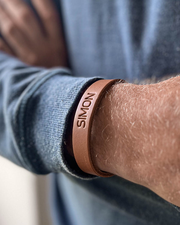 MONO skinnarmbånd i cognac skinn med navnet "Simon" gravert, båret rundt håndleddet på mann i blå strikk