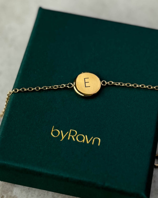 IBI LETTER armbånd i gullbelagt stål med bokstaven "E" gravert, liggende på mørkegrønn byRavn smykkeeske