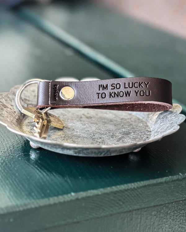 BOND nøkkelring i mørkebrunt skinn med graveringen "I'm so lucky to know you" og nøkkel i sinkfat på grønt bord