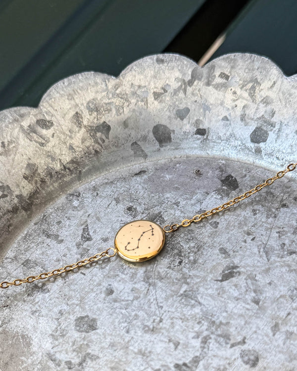 IBI STAR armbånd i gullbelagt stål med stjernebildet Skorpionen gravert på rund plate, liggende på sinkfat