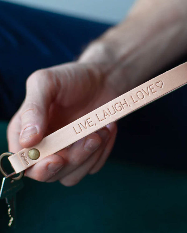 LUX nøkkelring i naturfarget skinn med messingnagle og personlig gravering "Live, laugh, love" ♡, holdt i hånden med nøkkel