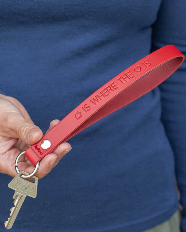 LUX nøkkelring i rødt skinn med graveringen "Home is where the heart is" skrevet med hus- og hjertesymbol, holdt i hånden