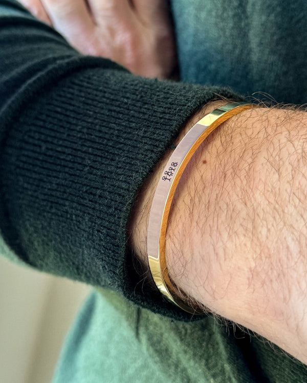 MARS STICK armbånd i gullbelagt stål med gravert fyrstikkfamilie, båret rundt håndleddet til mørkegrønn strikk