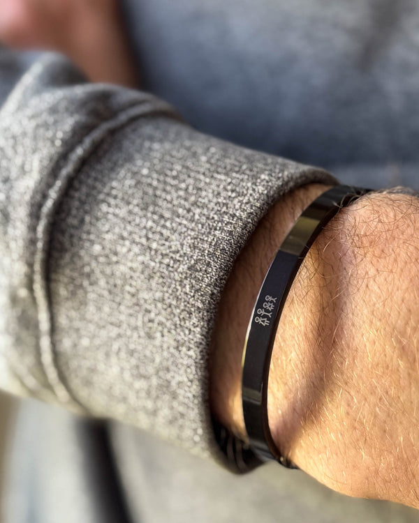 MARS STICK armbånd i svart stål med gravert fyrstikkfamilie, båret rundt håndleddet til grå strikk