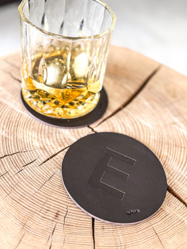 MOKKA glassbrikke i mørkebrunt skinn preget med bokstaven "E" på treskive ved whiskyglass på glassbrikke