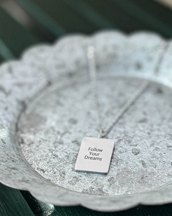 RAZ halskjede i sølvfarget rustfritt stål med graveringen "Follow Your Dreams" på rektangulær plate, på hvitpatinert sinkfat