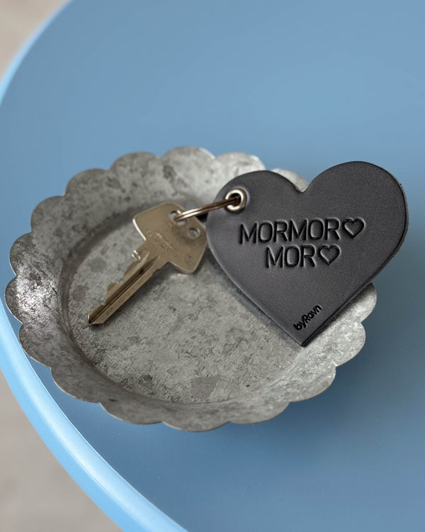 ROSE hjertenøkkelring i svart skinn med graveringen "Mormor ♡ Mor ♡" og nøkkel i sinkskål på blått bord