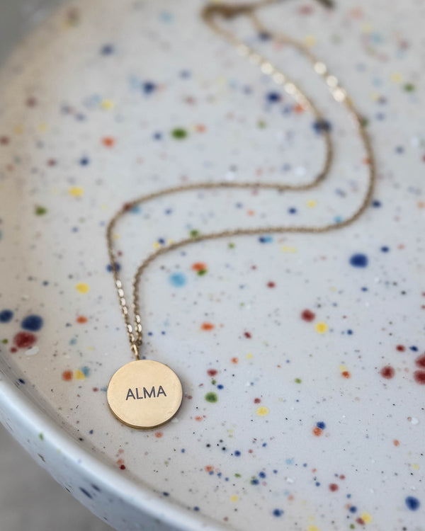 SIA NOTE navnehalskjede i gullbelagt stål med navnet "Alma" gravert på rundt anheng, på prikkete keramikkfat