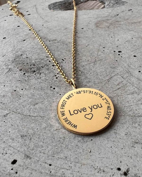 SIA CIRCLE halskjede i gullbelagt stål med "Love you" ♡ og "Where we first met" med koordinater gravert i sirkel, på betong