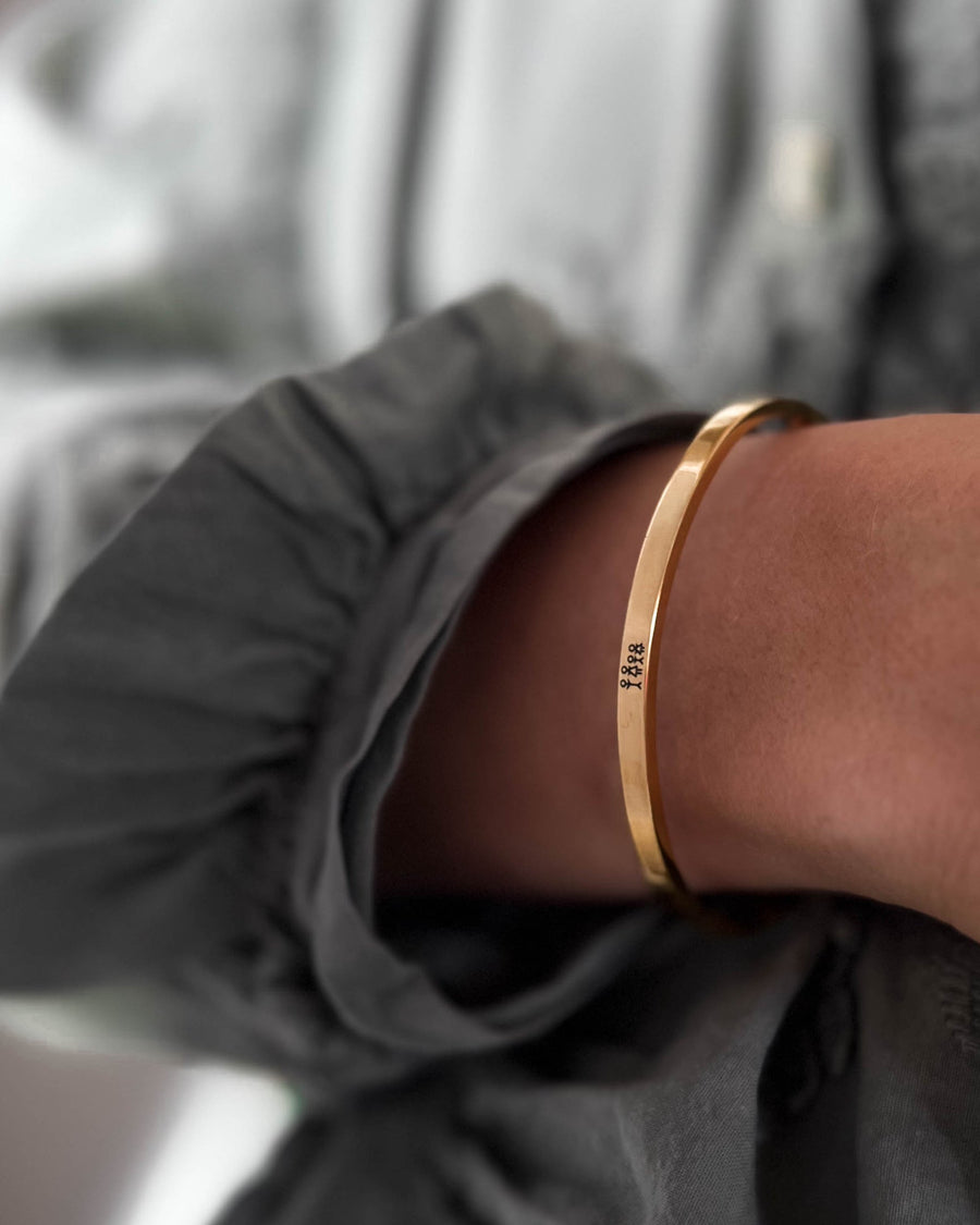 VENUS STICK armbånd i gullbelagt stål med gravert fyrstikkfamilie, båret rundt håndleddet til grå bluse