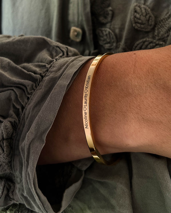 VENUS armbånd i gullbelagt stål med graveringen "Nicoline ♡ Laurits ♡ Kristian" rundt håndleddet, grå bluse