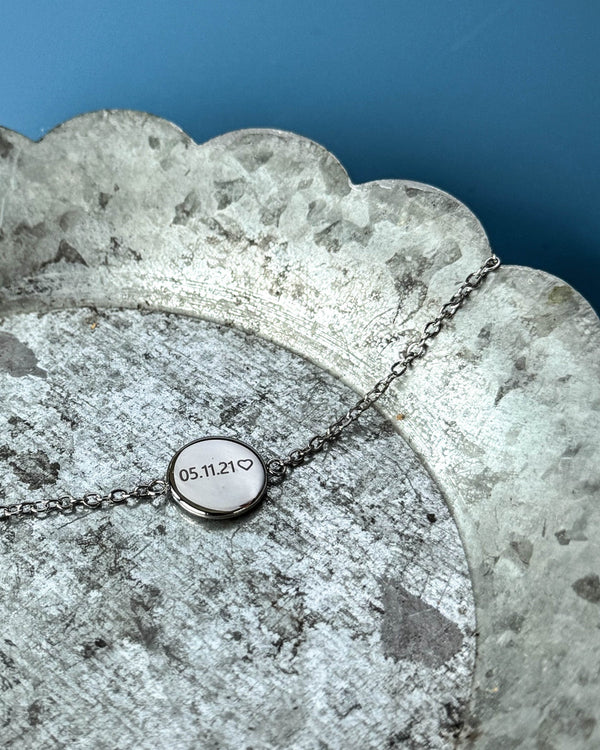 IBI NOTE armbånd i sølvfarget rustfritt stål med "05.11.21" ♡ gravert på rund plate, på sinkfat mot blå bakgrunn