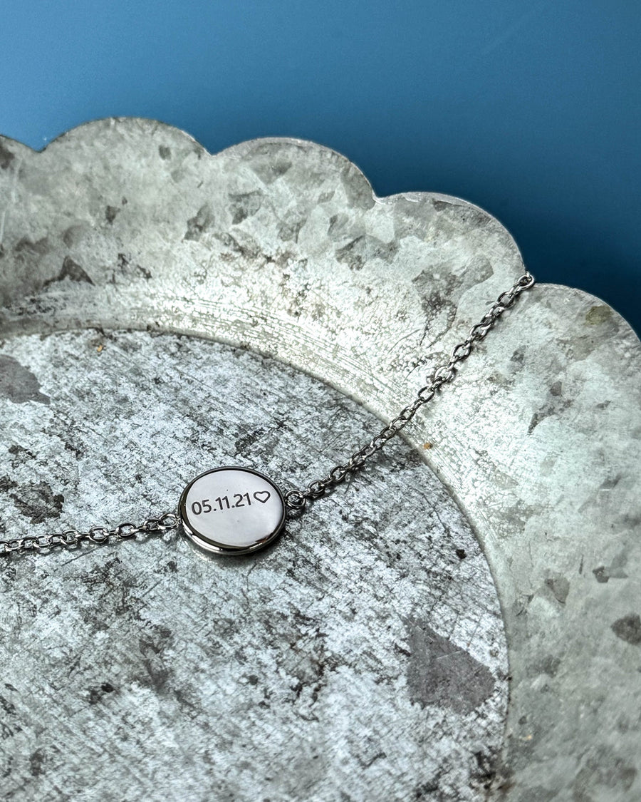 IBI NOTE armbånd i sølvfarget rustfritt stål med "05.11.21" ♡ gravert på rund plate, på sinkfat mot blå bakgrunn