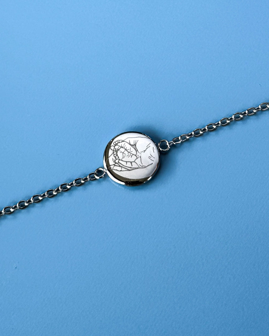 IBI PORTRAIT armbånd i sølvfarget rustfritt stål med strektegning av hender gravert på rund plate, mot blå bakgrunn