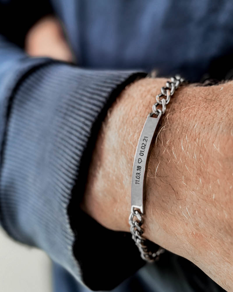 ARA armbånd i sølvfarget rustfritt stål med datoene "11.03.18 ♡ 01.02.21" gravert, rundt et mannshåndledd, blå sweatshirt