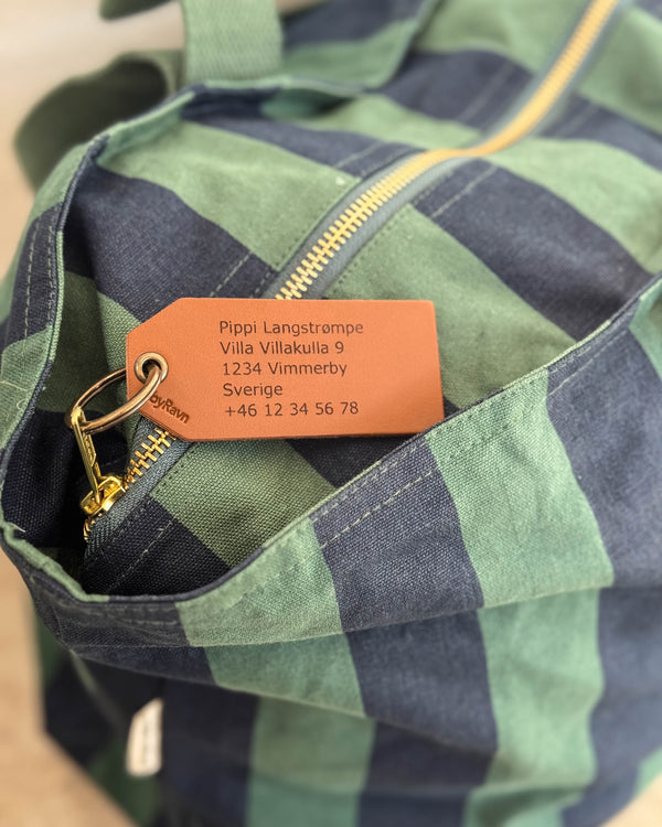 ATLAS bagasjelapp i skinn, her i cognac, med gravert adresse "Pippi Langstrømpe, Villa Villakulla 9" på grønn-blå stripete veske