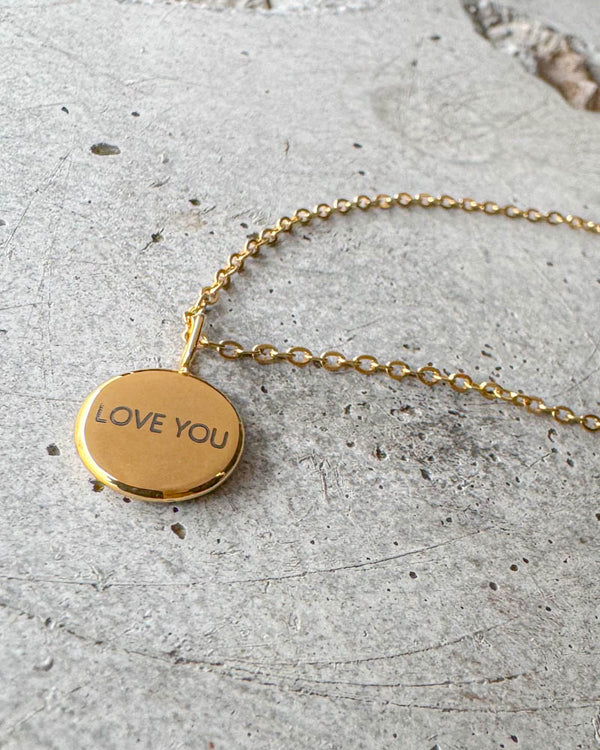 AYA NOTE halskjede i gullbelagt stål med graveringen "Love you" på lite rundt anheng, liggende på betong