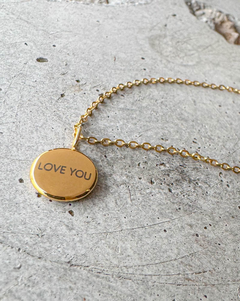 AYA NOTE halskjede i gullbelagt stål med graveringen "Love you" på lite rundt anheng, liggende på betong
