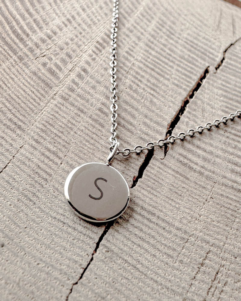 AYA LETTER halskjede i sølvfarget rustfritt stål med bokstaven "S" gravert på lite rundt anheng, på lyst tre med sprekk
