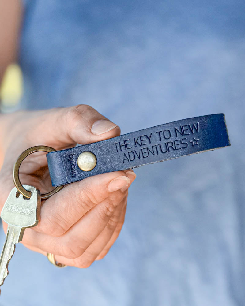 BOND nøkkelring i mørkeblått skinn med personlig gravering "The key to new adventures" ☆, holdt i hånden med nøkkel