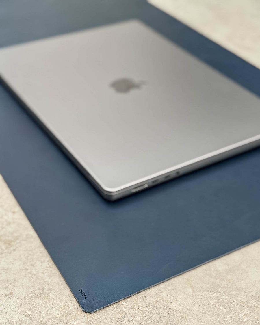XXL DESK skriveunderlag i mørkeblått kjerneskinn med byRavn-preg under laptop på lys bordplate