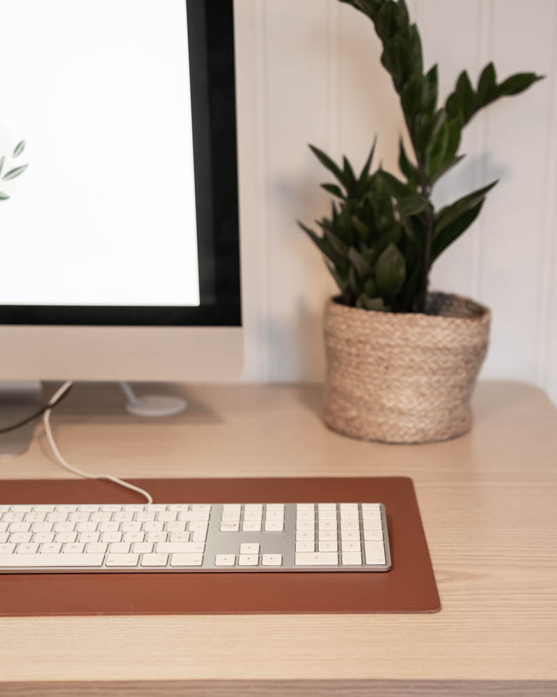 XXL DESK skriveunderlag i cognac skinn under tastatur foran iMac med grønn plante i flettet kurv