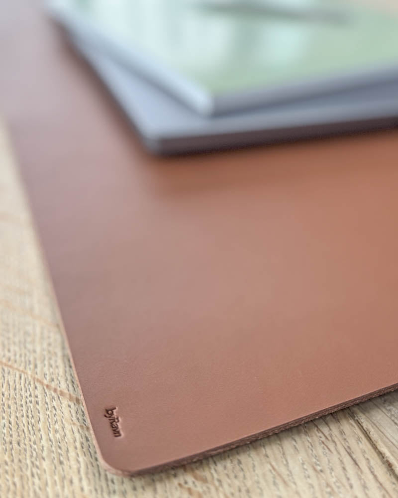 XXL DESK skriveunderlag i cognac kjerneskinn, nærbilde av hjørnet med byRavn-preg under laptop