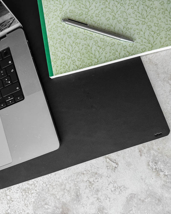 DESK skriveunderlag i svart skinn sett ovenfra med laptop, blomstrete notatbok og kulepenn på lys bordplate