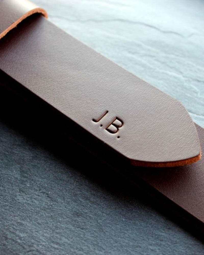 EDGE belte i mørkebrunt skinn sett på nært hold med initialene "J.B." gravert på tuppen, på skiferplate