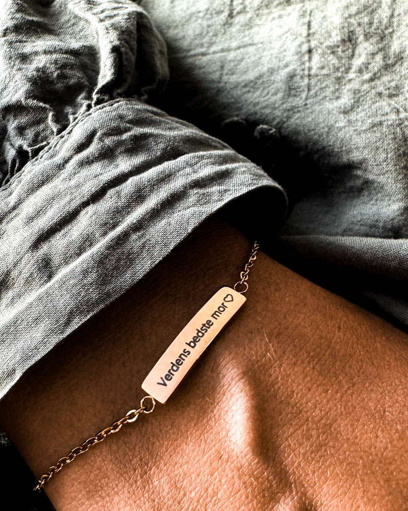 EVI armbånd i gullbelagt stål med graveringen "Verdens bedste mor" ♡ rundt håndleddet, grå linskjorte