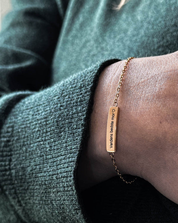 EVI armbånd i gullbelagt stål med "Verdens bedste mor" ♡ gravert på avlang plate, båret rundt håndleddet til grønn strikk