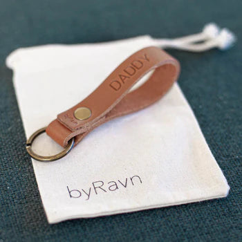 Gavepose i hvitt stoff med byRavn-logo og nøkkelring i cognac skinn med graveringen "Daddy" på grønt underlag