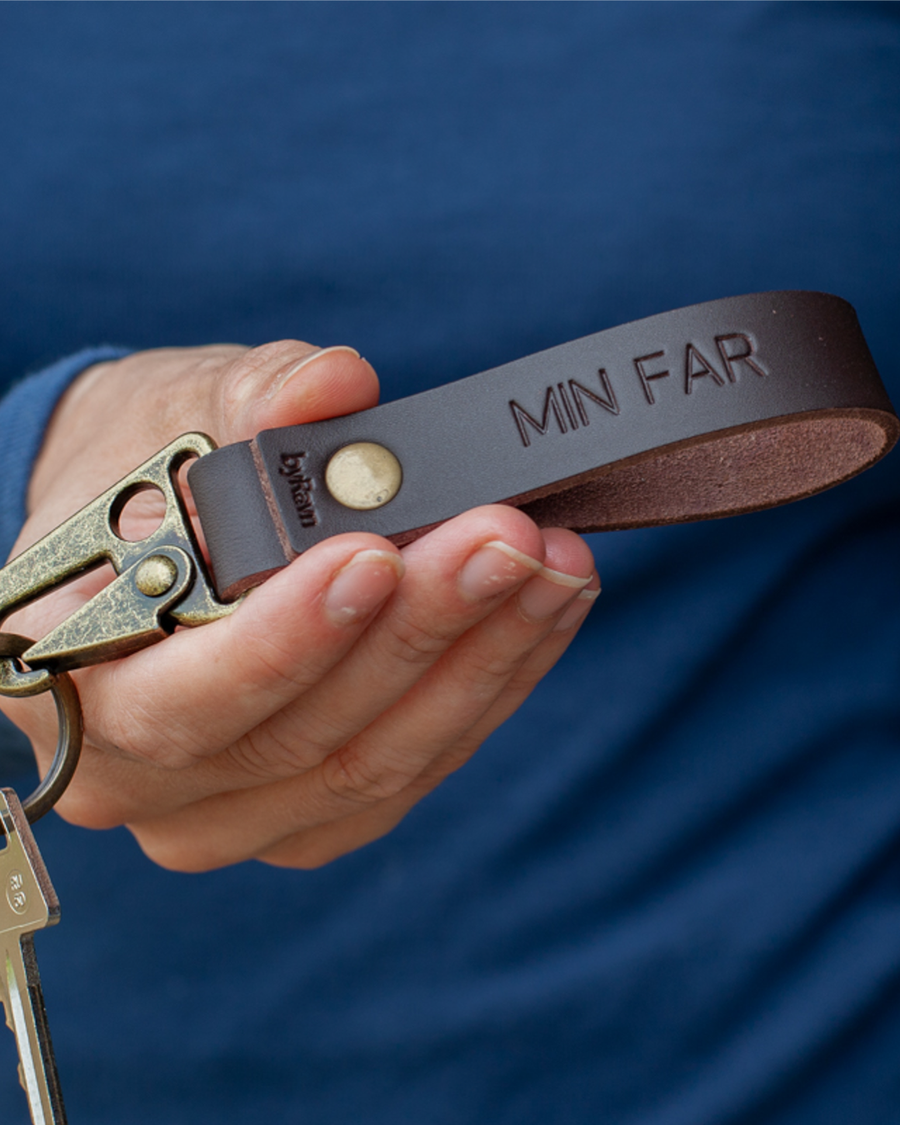 HOOK nøkkelring i mørkebrunt skinn med karabinkrok i antikk messing og graveringen "Min far", holdt i hånden med nøkkel