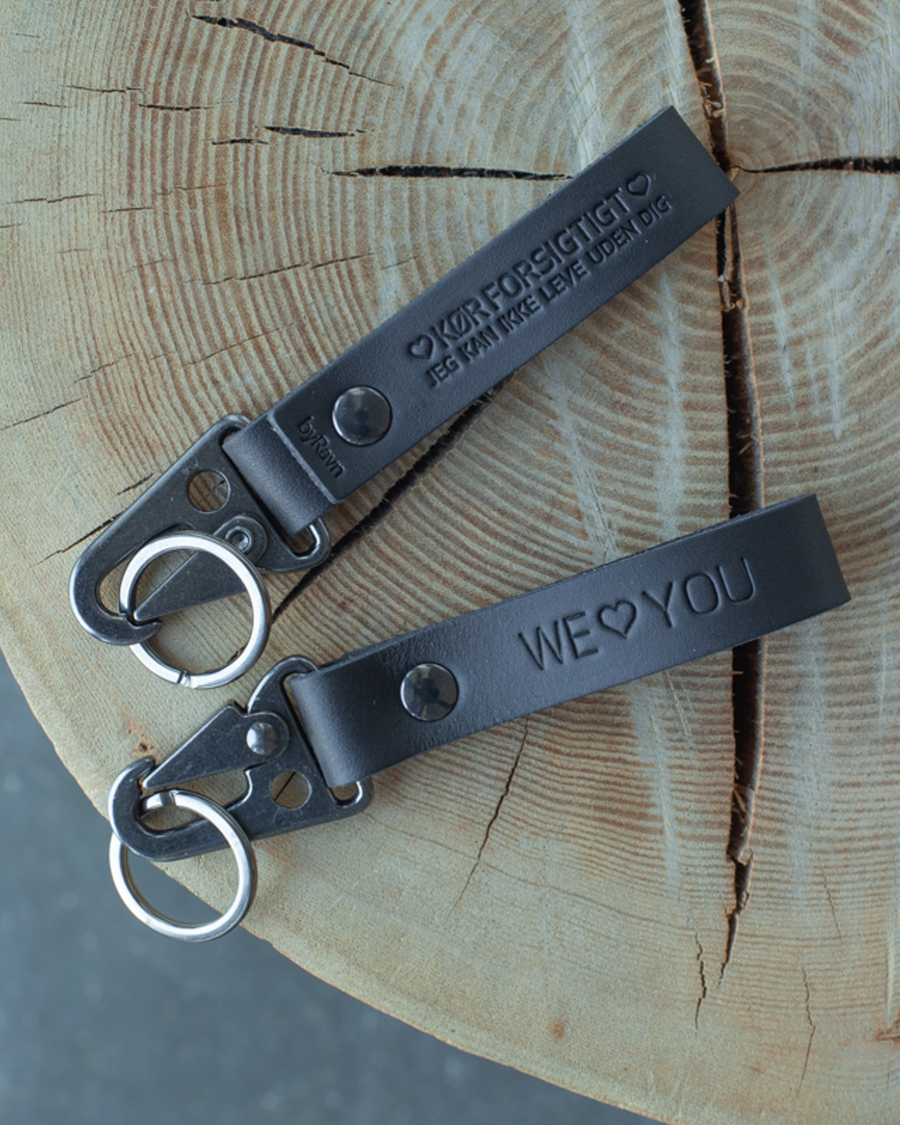 To HOOK nøkkelringer i svart skinn på trestubbe med graveringene "We ♡ you" og "Kør forsigtigt – jeg kan ikke leve uden dig"