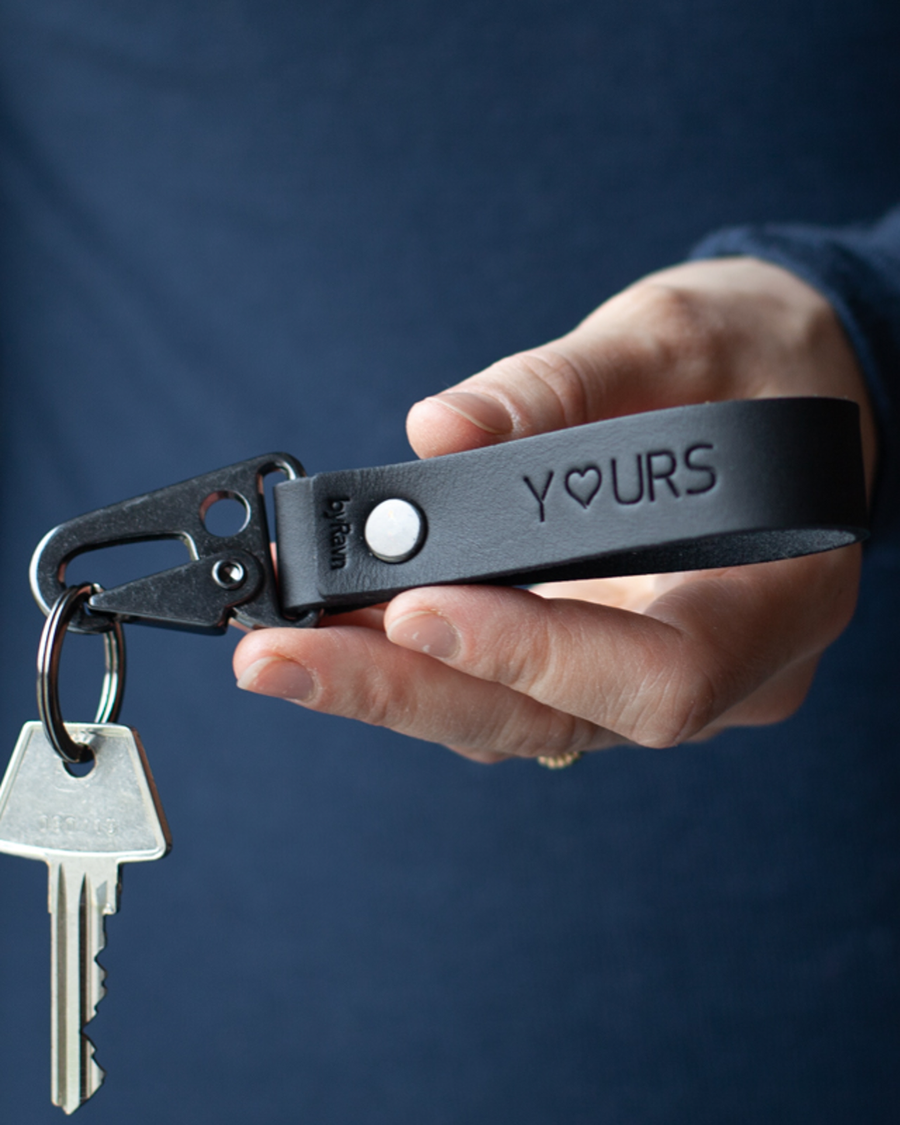HOOK nøkkelring i svart skinn med graveringen "Y♡urs" og karabinkrok i svart stål, holdt i hånden med nøkkel