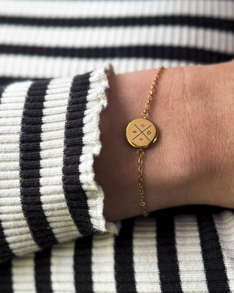 IBI X armbånd i gullbelagt stål med kryss, bokstaver og hjerte gravert, båret rundt håndleddet til stripete bluse