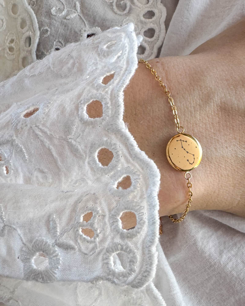 IBI STAR armbånd i gullbelagt stål med stjernebilde gravert på rund plate, båret rundt håndleddet til hvit blondebluse