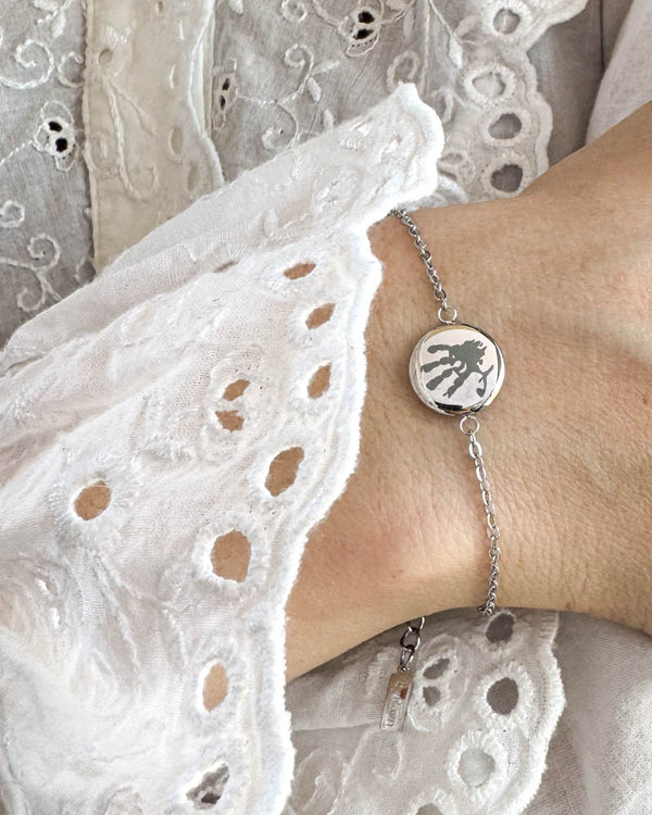 IBI MOTIF armbånd i sølvfarget rustfritt stål med gravert håndavtrykk, båret rundt håndleddet til hvit blondebluse