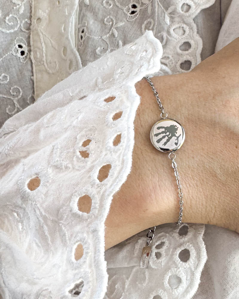 IBI MOTIF armbånd i sølvfarget rustfritt stål med gravert håndavtrykk, båret rundt håndleddet til hvit blondebluse