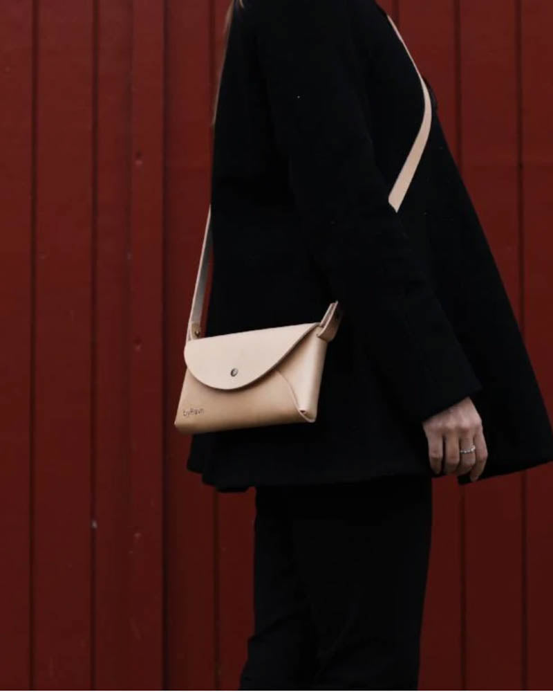 INA skulderveske i naturfarget skinn båret crossbody av kvinne i svarte klær foran rød trekledning