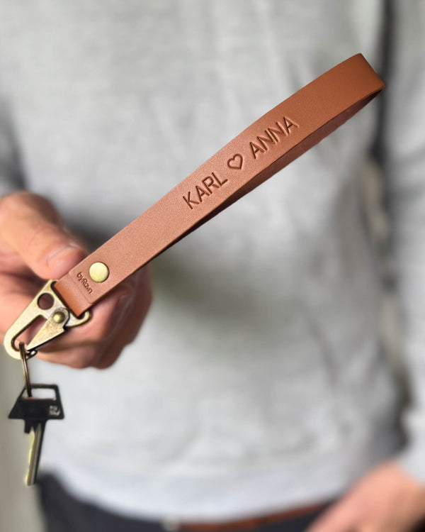 IRON nøkkelring i cognac skinn med navnene "Karl ♡ Anna" gravert, holdt i hånden med nøkkel