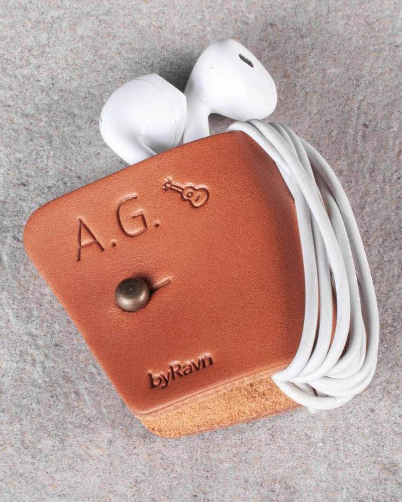 KAX hodetelefonholder i cognac skinn med initialene "A.G." og gitar gravert, med earpods på grå stein