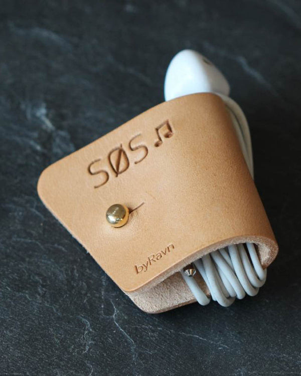 KAX hodetelefonholder i naturfarget skinn med graveringen "Søs" ♪ og messingknapp, med earpods på mørk skifer