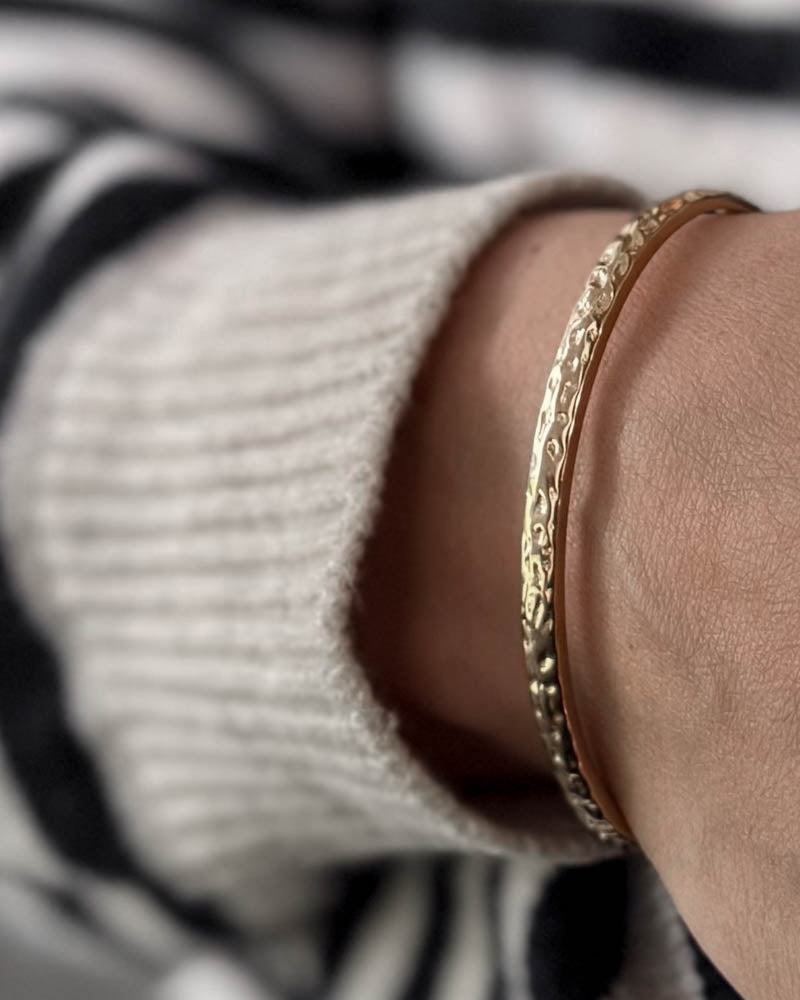 LUNA armbånd i gullbelagt stål med hamret overflate rundt håndleddet, hvit strikk og stripete bluse