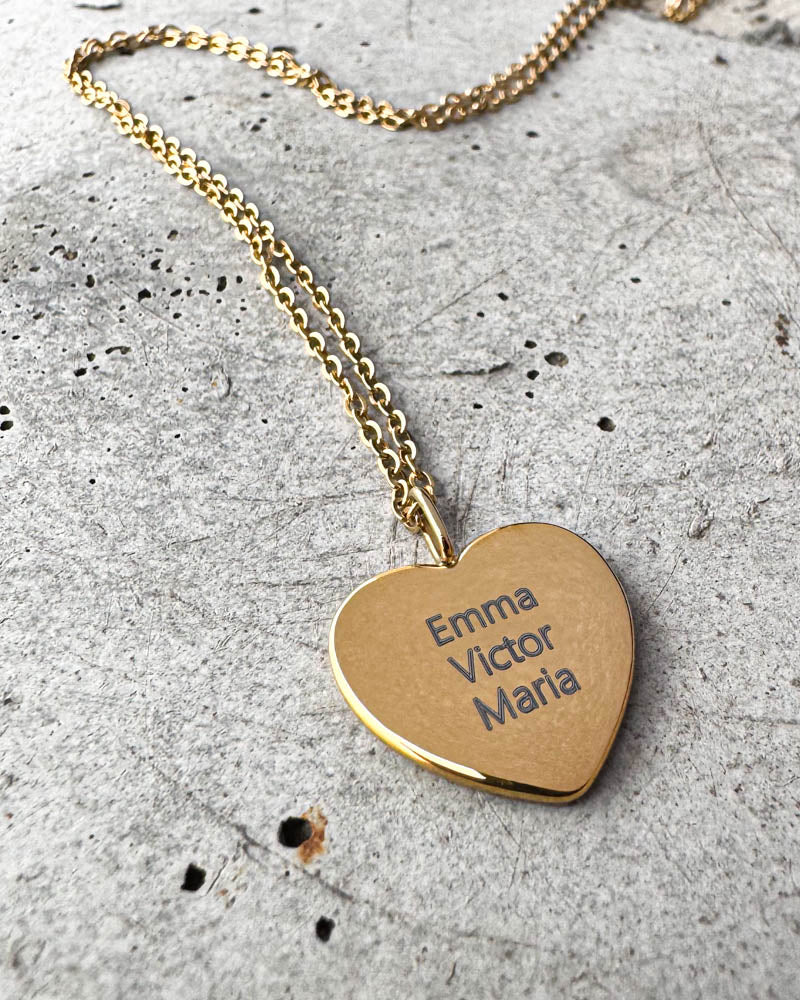 LUV QUOTE navnehalskjede i gullbelagt stål med navnene "Emma Victor Maria" gravert på hjerteanheng, liggende på betong