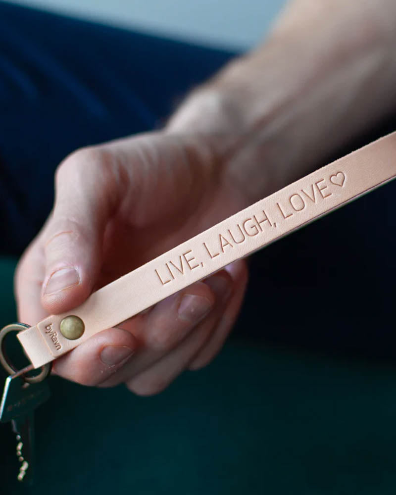 LUX nøkkelring i naturfarget skinn med messingnagle og personlig gravering "Live, laugh, love" ♡, holdt i hånden med nøkkel