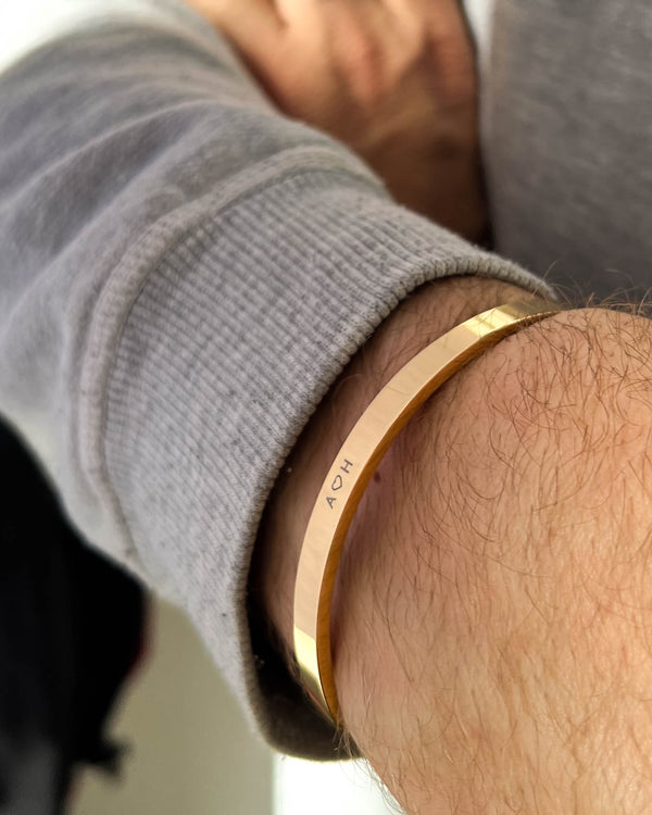 MARS armbånd i gullbelagt stål med initialene "A ♡ H" gravert rundt mannshåndledd, grå collegegenser