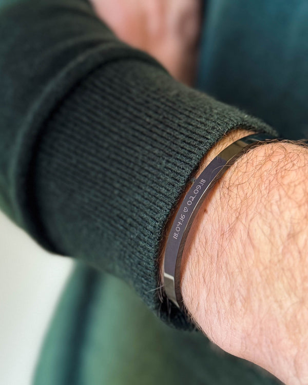 MARS SCRIPT armbånd i svart stål med datoene "18.04.16 ♡ 02.09.18" gravert i håndskrift, båret til mørkegrønn strikk