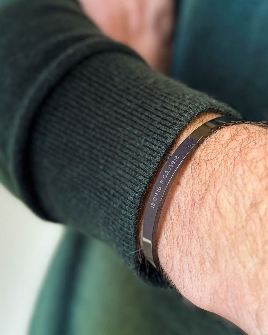 MARS SCRIPT armbånd i svart stål med datoene "18.04.16 ♡ 02.09.18" gravert i håndskrift, båret til mørkegrønn strikk