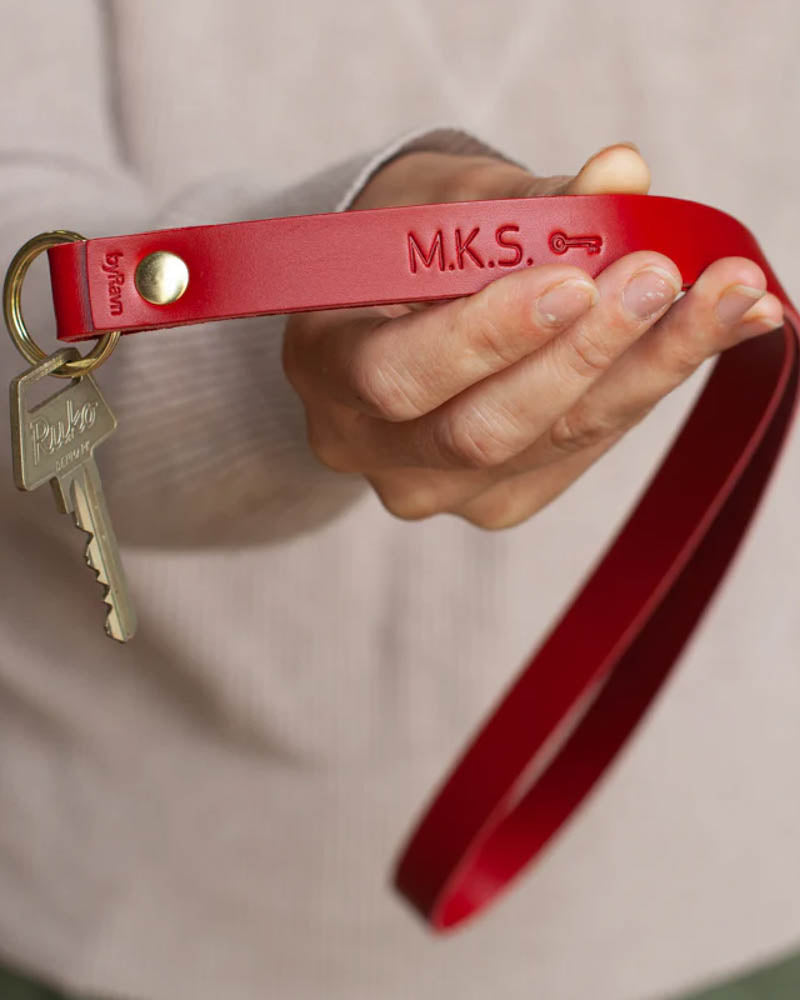MAX nøkkelring i rødt skinn som lang stropp med initialene "M.K.S." og nøkkelsymbol gravert, holdt i hånden med nøkkel
